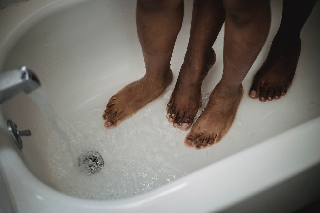 Two masculine appearing darker toned feet standing intimately in a bathtub filling with water. Photo is cropped to show only beneath the knee cap. 