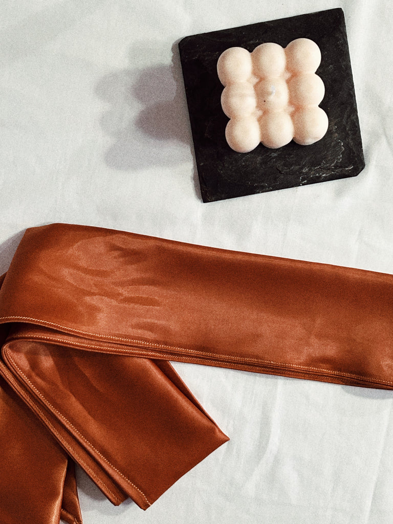 Copper/ Bronze satin blindfold for intimacy, sexual wellness and pleasure tied with large bow. Blindfold is pictured close up neatly folded on white sheets. Alchemy wellness co blindfold is and intended to inspire sexual exploration and for bondage or sensory play for adults. 