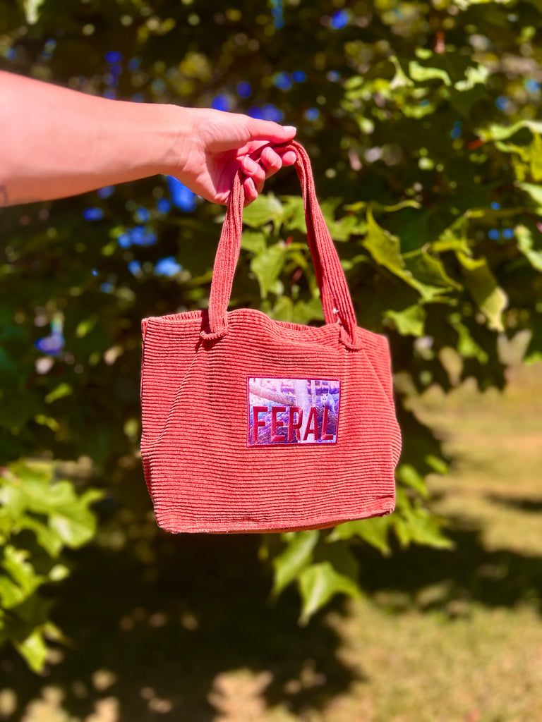 Red corduroy reworked/repurposed/upcycled bag held by a hand with a blurred green leafy background