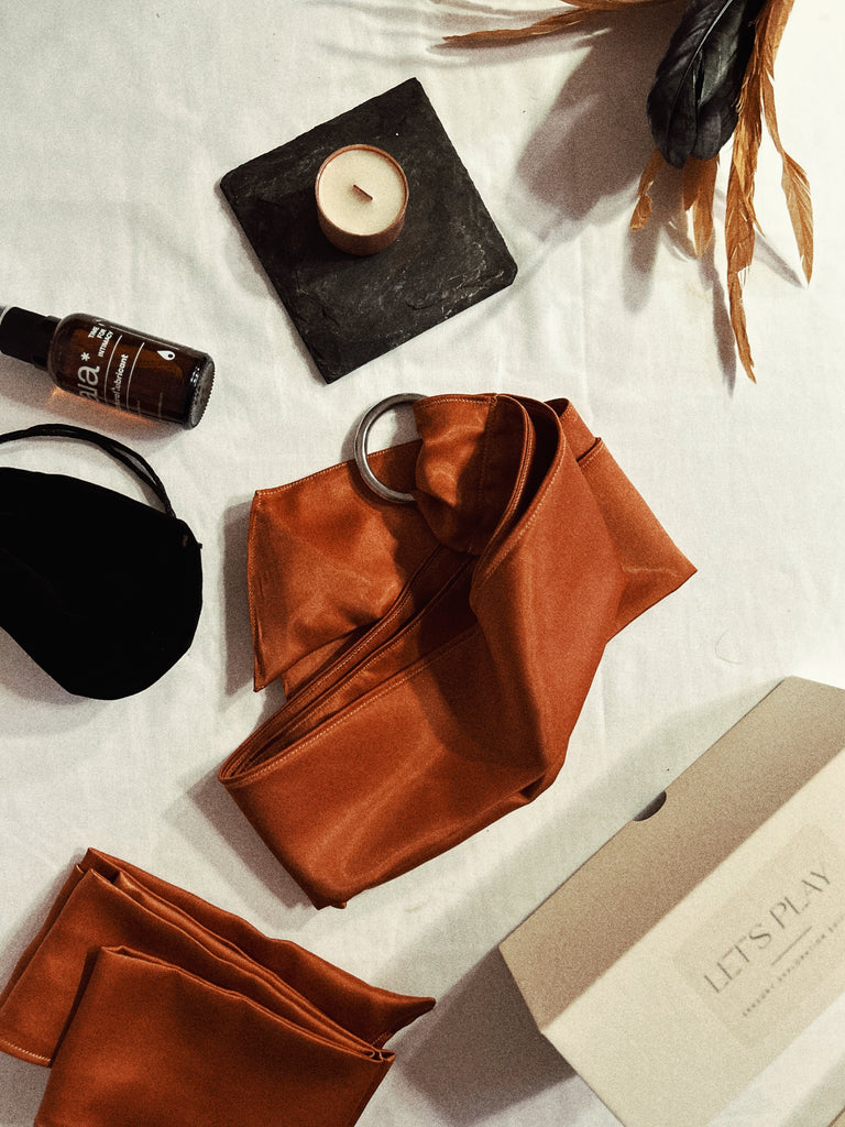 Copper/ Bronze satin restraint ties for intimacy, sexual wellness and pleasure placed in centre with contents of product bundle scattered around. Copper satin Blindfold is pictured laid on white sheets and intended for bondage or sensory play for adults. A velvet pouch, travel size water based natural lube, feathers and product box surround restraints. 