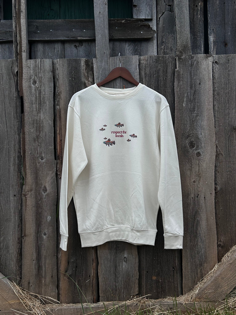 Cream crew neck sweater with multi indigenous salmon design and text in maroon with the saying ‘respect the logo’ in the centre. A nature, sustainable designed sweater hanging against a wooden fence. 