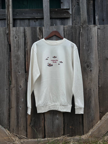 Cream crew neck sweater with multi indigenous salmon design and text in maroon with the saying ‘respect the logo’ in the centre. A nature, sustainable designed sweater hanging against a wooden fence. 