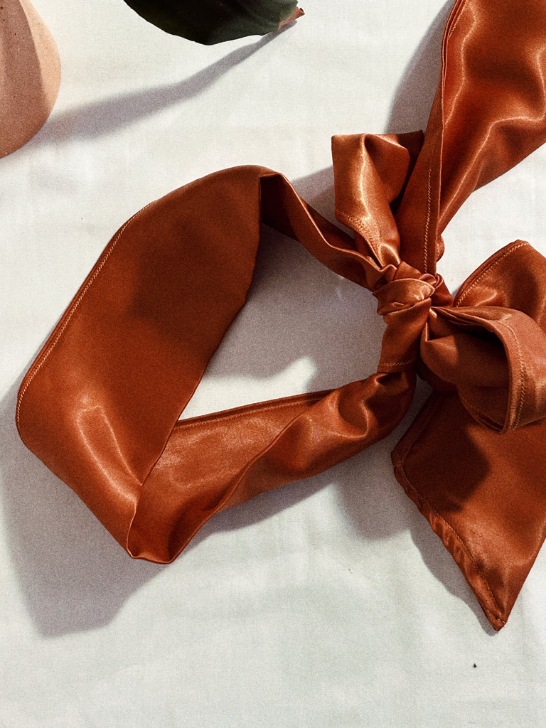 Copper/ Bronze satin blindfold for intimacy, sexual wellness and pleasure tied with large bow. Blindfold is pictured up close laid on white sheets and intended for bondage or sensory play for adults. 