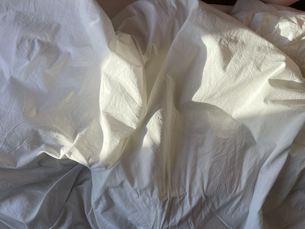 Close up image of rustled white bed sheets with light streaming in over the top half of the frame. 