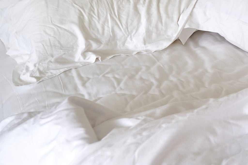 Close up photo of bright white bed sheets with blankets on lower half of page and the corner of a pillow in the right corner. 