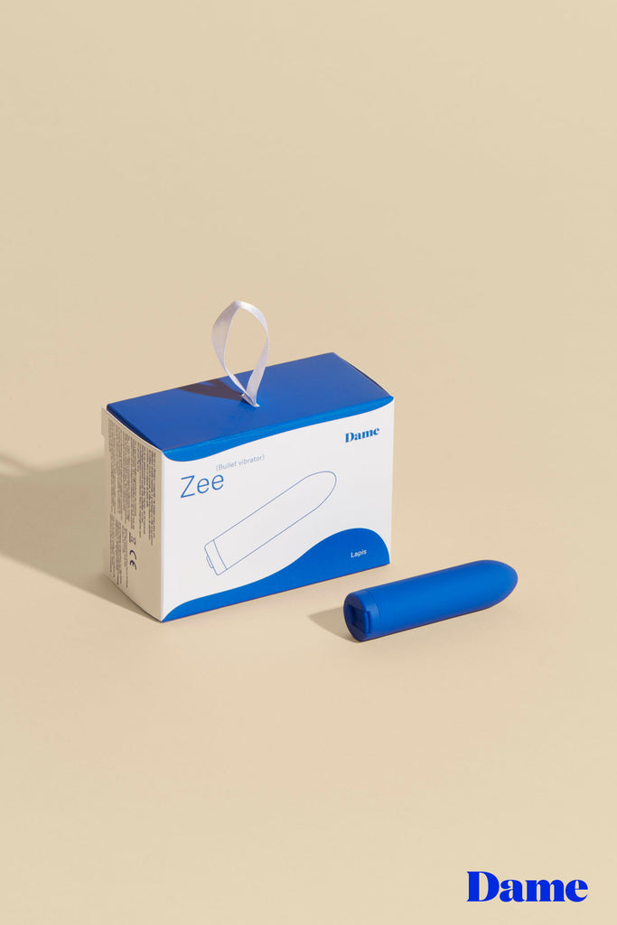 Blue Zee bullet vibe by Dame displayed laying horizontally on solid beige background with product box featured behind it. 