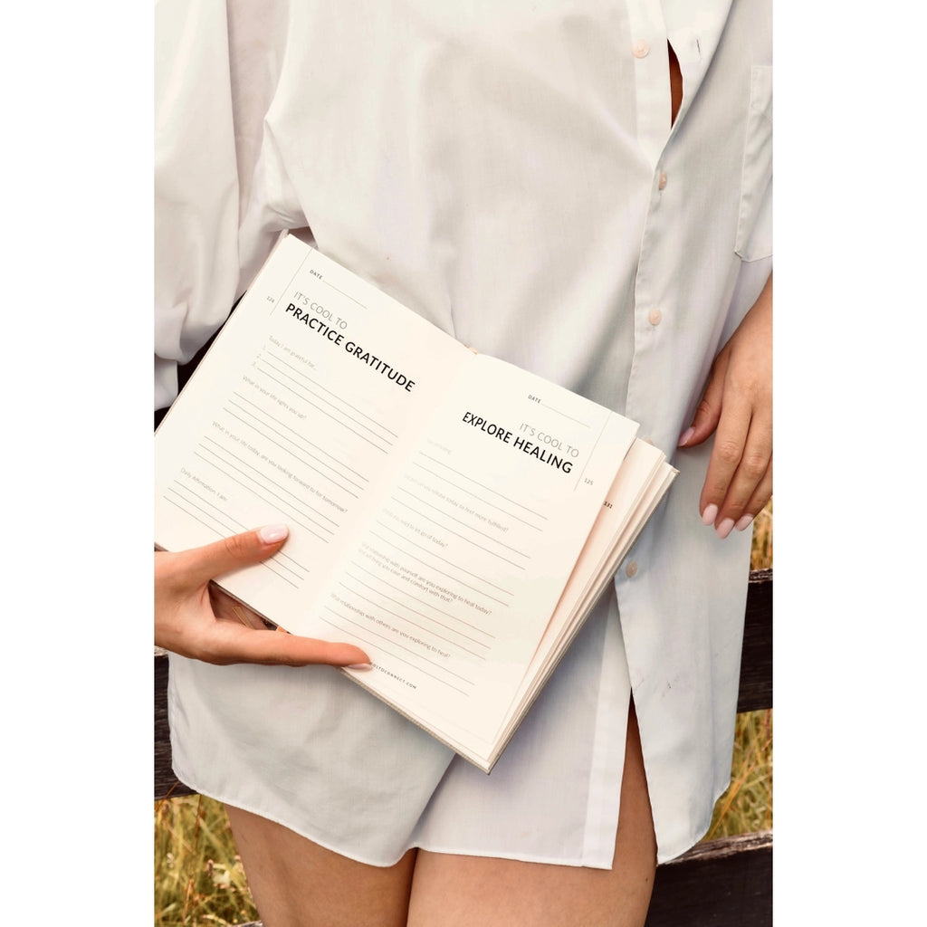 Woman in white button down shirt holding cool to connect journal open displaying text prompts on two of the pages. 