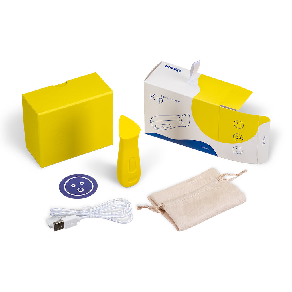 Prism shaped intimate massage Kip in lemon by Dame displayed standing vertically centered around product box contents including carying case, charging cable and sticker on solid white background.