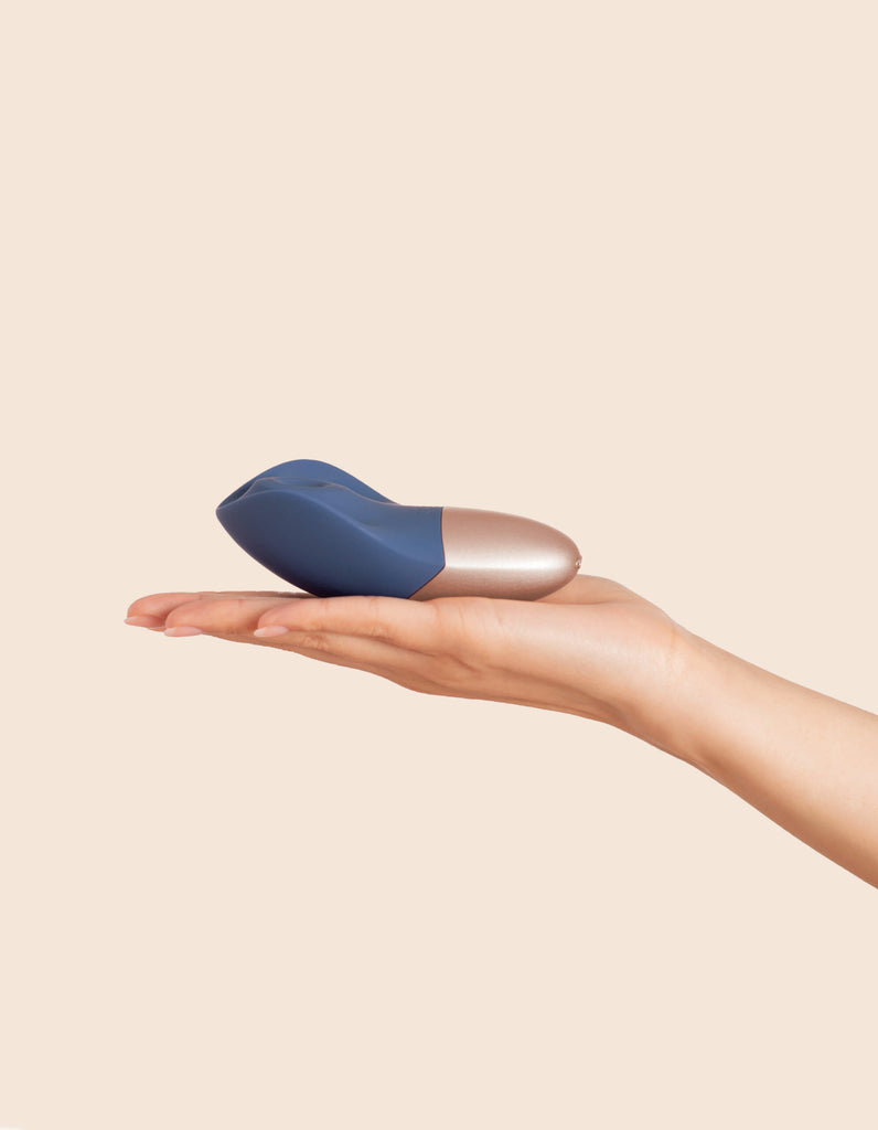 Feminine hand holding Deia Arouser clitoral stimulator laying in palm in front of beige background.