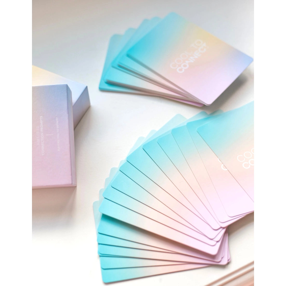 Product display of Cool to connect conversation cards for kids. Rainbow card deck split and white surface and spread out displaying pastel rainbow colours and cool to connect logo. Includes part of product box on left side. 