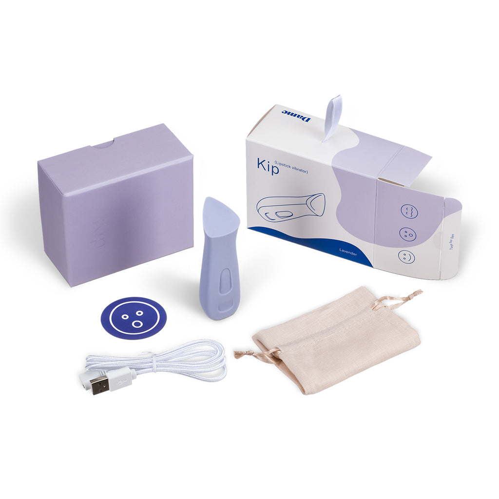 Prism shaped intimate massage Kip in lavender by Dame displayed standing vertically centered around product box contents including carying case, charging cable and sticker on solid white background.