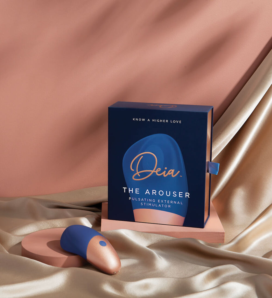Deia Arouser clitoral stimulator displayed laying on satin sheet in front of product box with solid pink background.