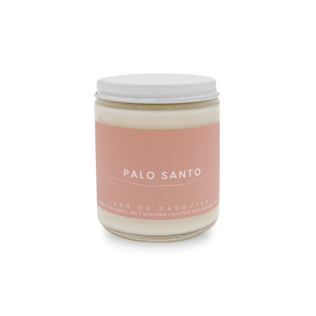 A handmade coconut soy wax candle by Land of Daughters in scent Palo Santo displayed on solid white background. Candle stands alone in center of image with pink coloured label.  