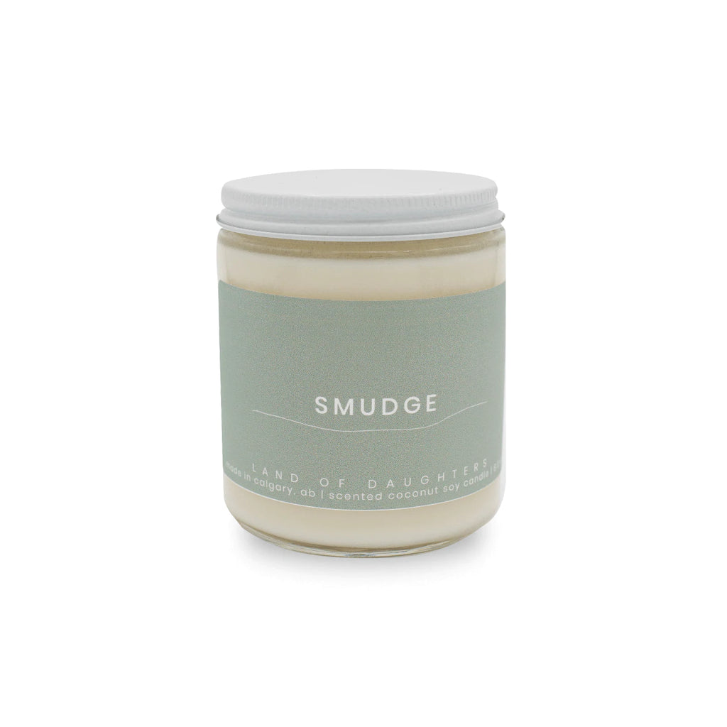 A handmade coconut soy wax candle by Land of Daughters in scent smudge displayed on solid white background. Candle stands alone in center of image with sage coloured label.  
