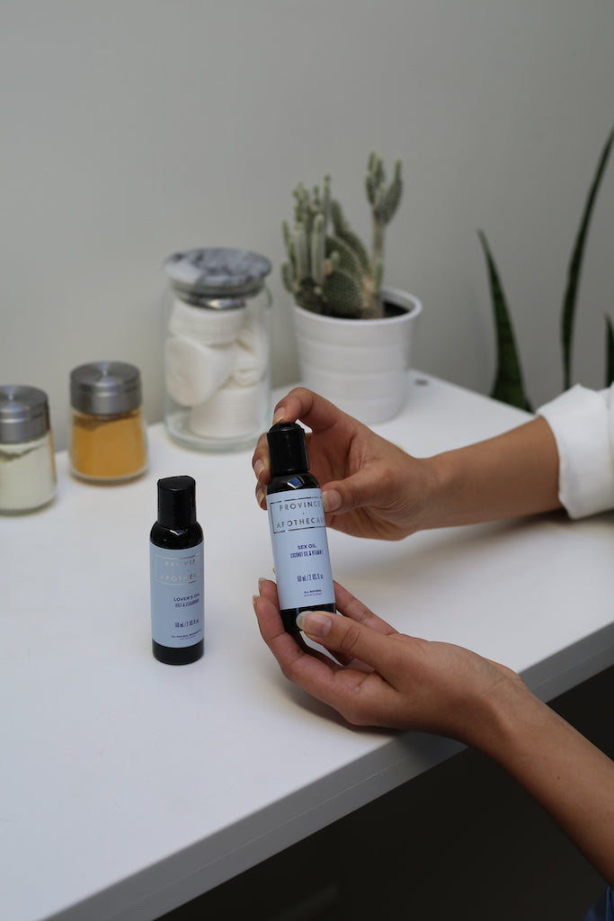 Province Apothecary all natural sexual wellbeing and intimacy care kit includes lovers oil and sex oil displayed in bathroom on solid white counter top with toiletry items in background. A Womans hands are pictured agains countertop holding and displaying 60ml bottle of sex oil. Naturally formulated from botanicals and plant derived ingredients made in Canada by women owned business 