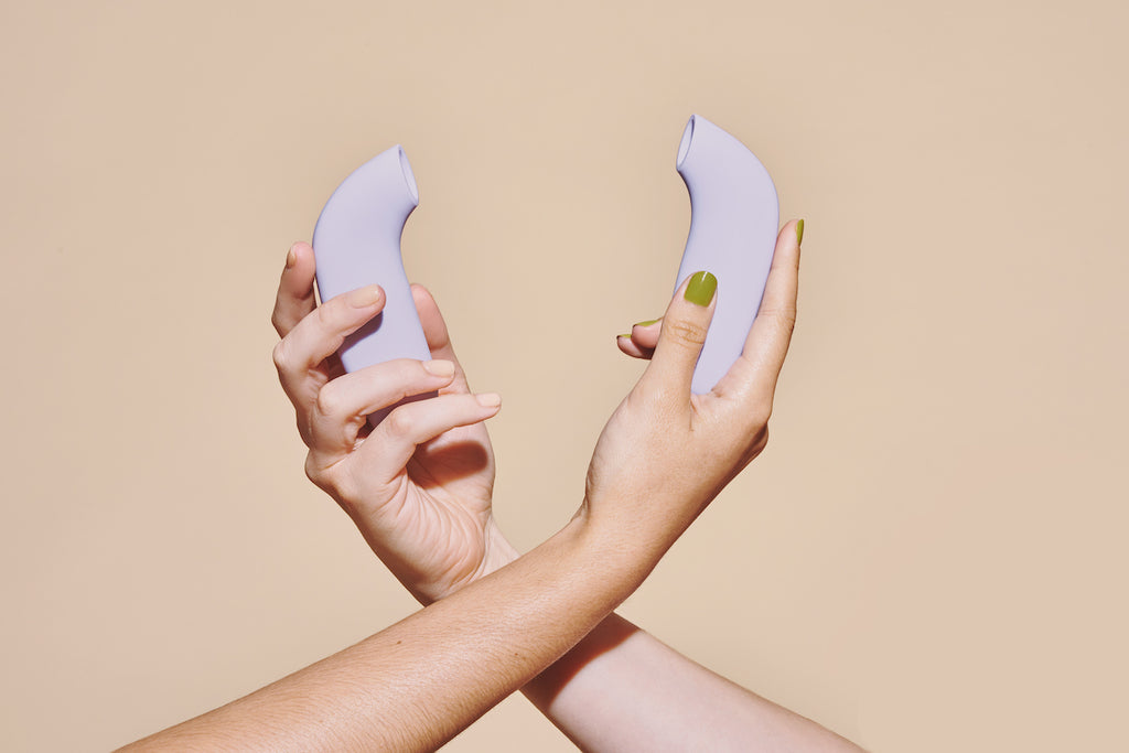 Two lavender coloured AER suction stimulators by dame held in crossed hands with products facing each other on solid beige background. 