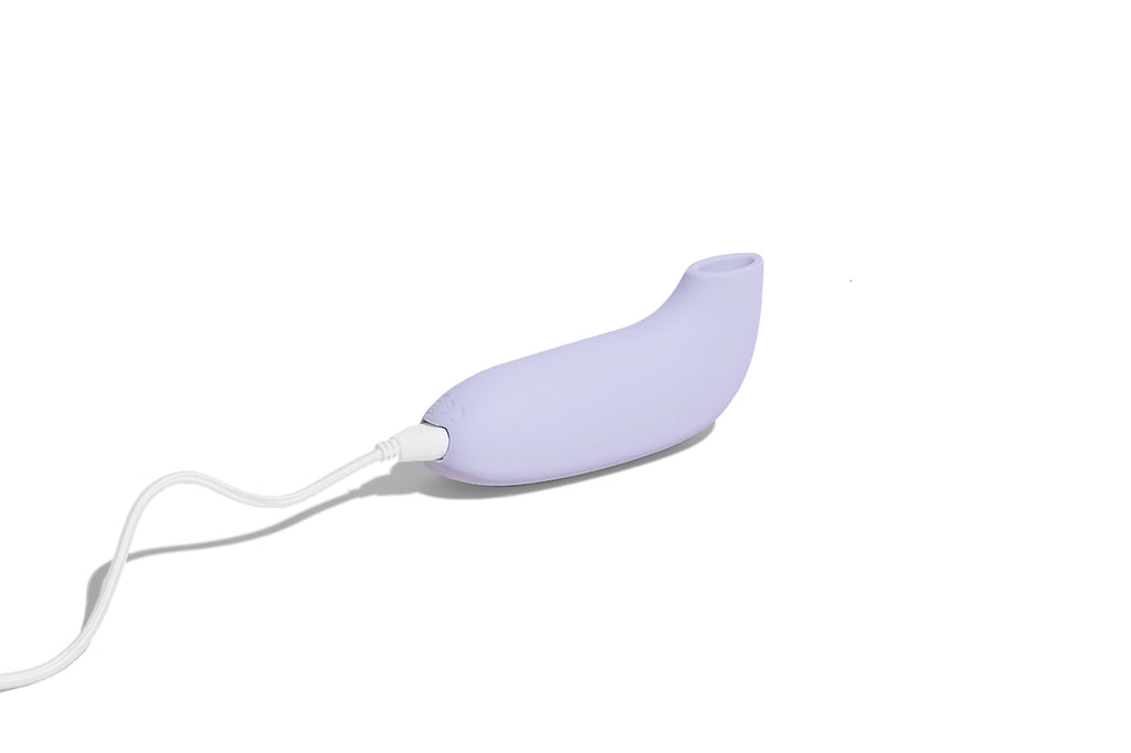 Lavender coloured AER by dame, suction stimulator displayed laying down with charging cable attached on solid white background.