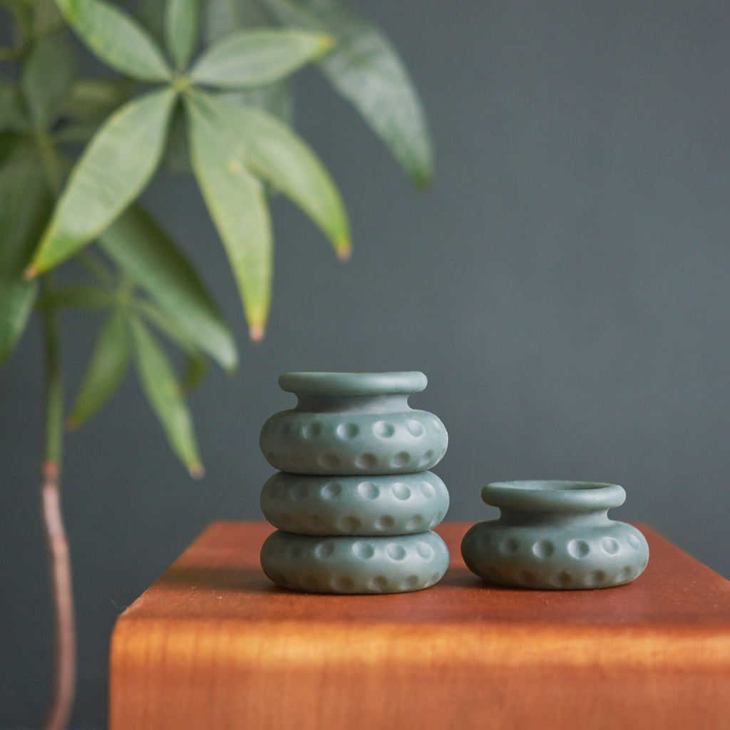 Oh Nut brand silicon intimate wellness buffer in the colour aloe being displayed on bedside table stacked on one another. A single buffer sits beside. The buffers are placed in front of large tree and dark green background