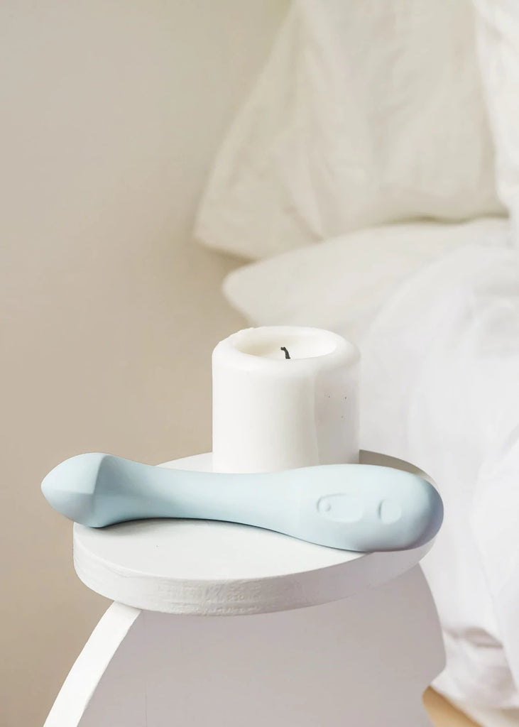 Light blue Dame personal massager Arc displayed in bedside scene laid horizontally on bedside table with large candle. 