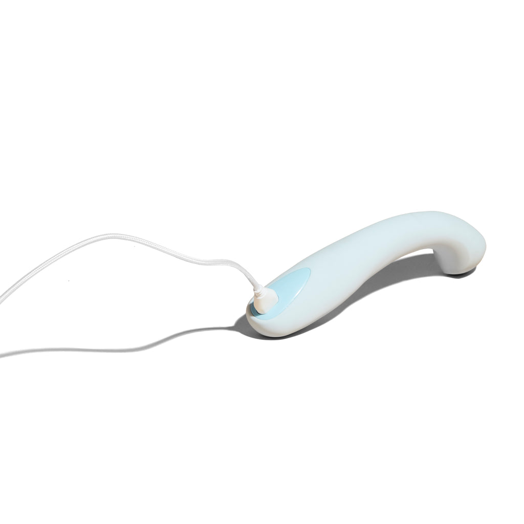 Light blue Dame personal massager Arc displayed on solid white background laid face down with charging cable attached.