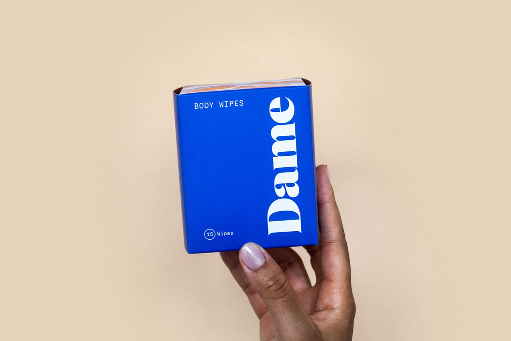 feminine hand holding blue box of dame body wipes in 15 count against solid beige background. 