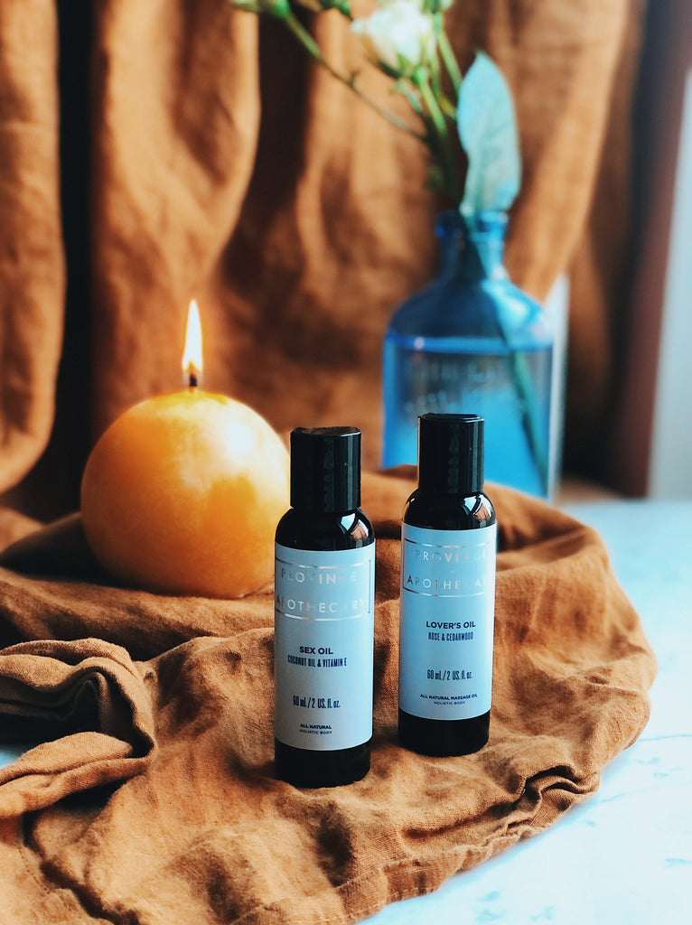 Provice apothecary's sexual wellbeing and intimacy care kit - lover oil and sex oil in 60ml bottles displayed in a romantically decorated scene featuring roses and orange glowing candle in background against rusty orange material. Bottles are placed beside each sitting on fabric with sustainable packaging, naturally derived ingredients that are effective and dermatologist recommended. Made in Canada by women owned company. 
