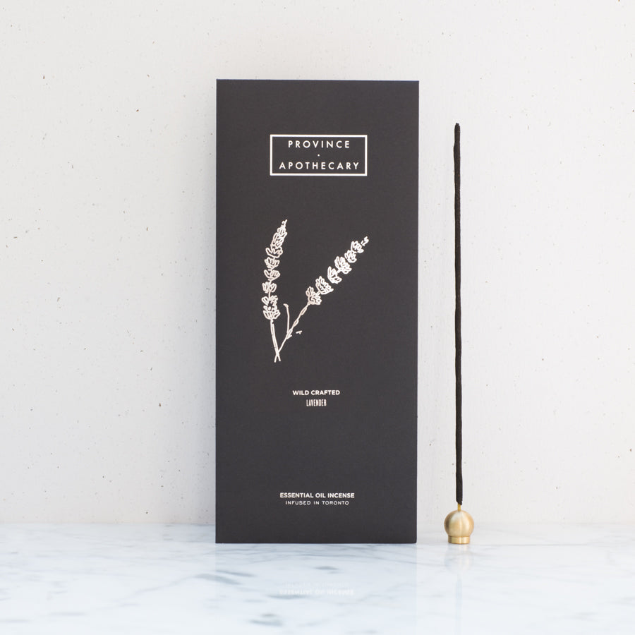Black foil decorated package of Provice apothecary incense in lavender displayed standing against white background. Province apothecary brass round lunar incense holder standing to the right of package holding single incense.