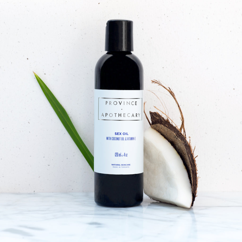 120ml black bottle of Province Apothecary sex oil being displayed against solid white background. Demonstrating natural ingredients with partial coconut flesh and shell standing to right of product bottle and long skinny piece of plant tucked behind.