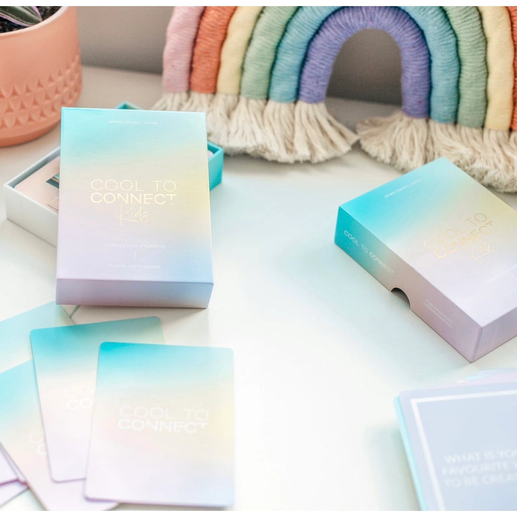 Lifestyle scene of cool to connect kids card deck placed on white surface with decorative fabric rainbow in background. Includes the deck of cards separated on table top surface displaying the pastel rainbow colours deck box with logo and cards spread out with logo and text.  
