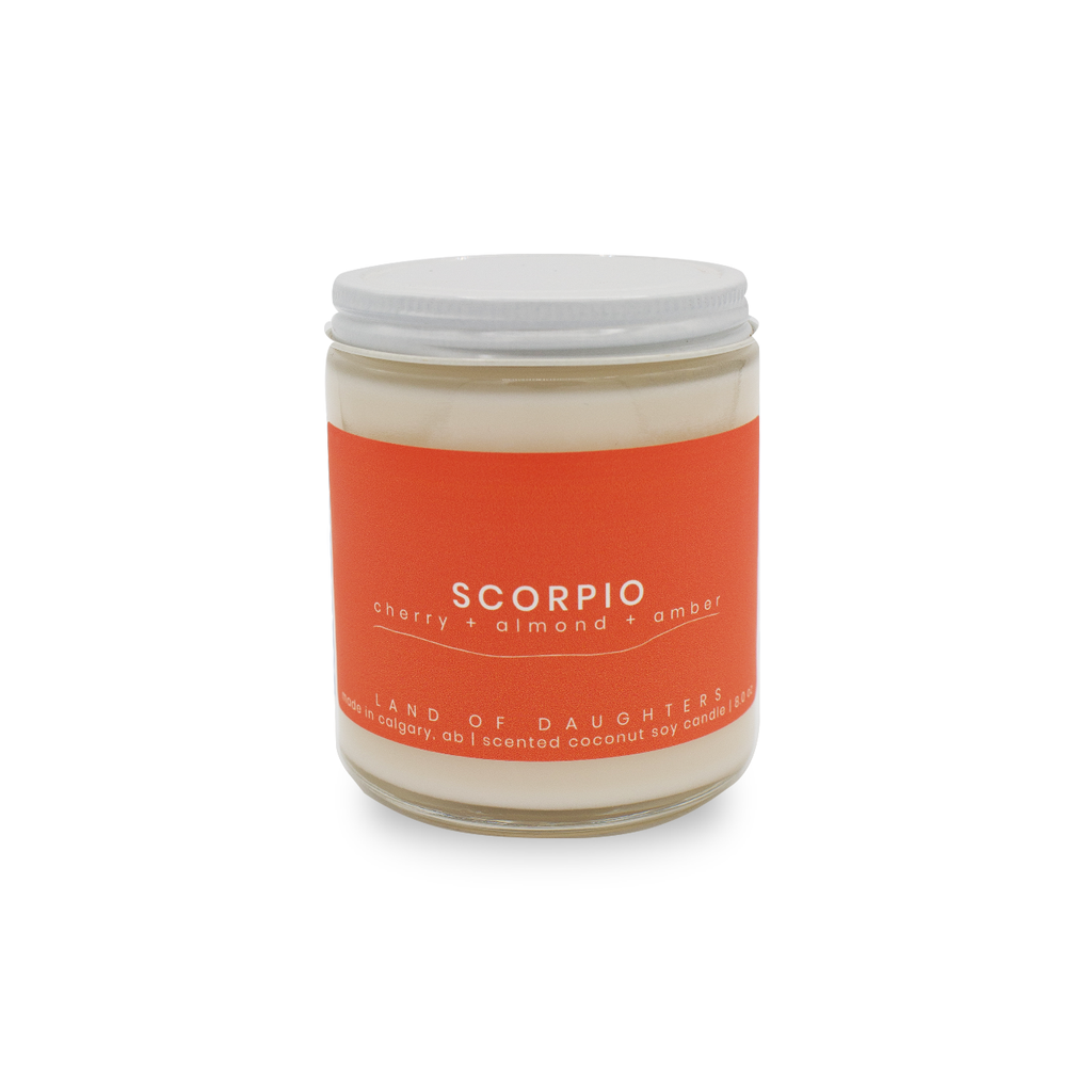 A handmade soy wax candle by Land of Daughters in scent scorpio displayed on solid white background. Candle stands alone in center of image with bold orange label.  