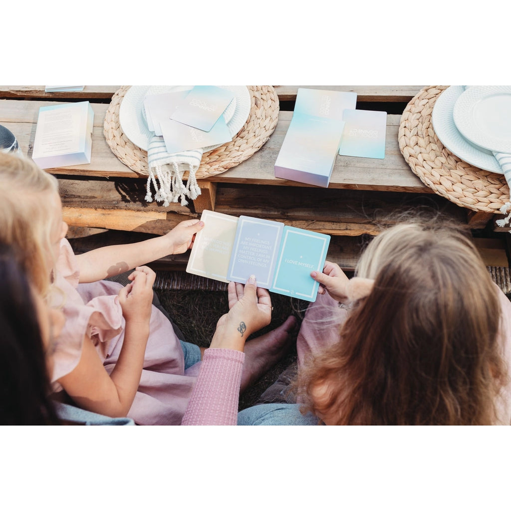 Lifestyle scene of cool to connect kids card deck being used by two children sitting at a wooden bench and picnic table set up. Each child holds cards in their hands while an adult oversees comparing the prompts  or text given on each card. 