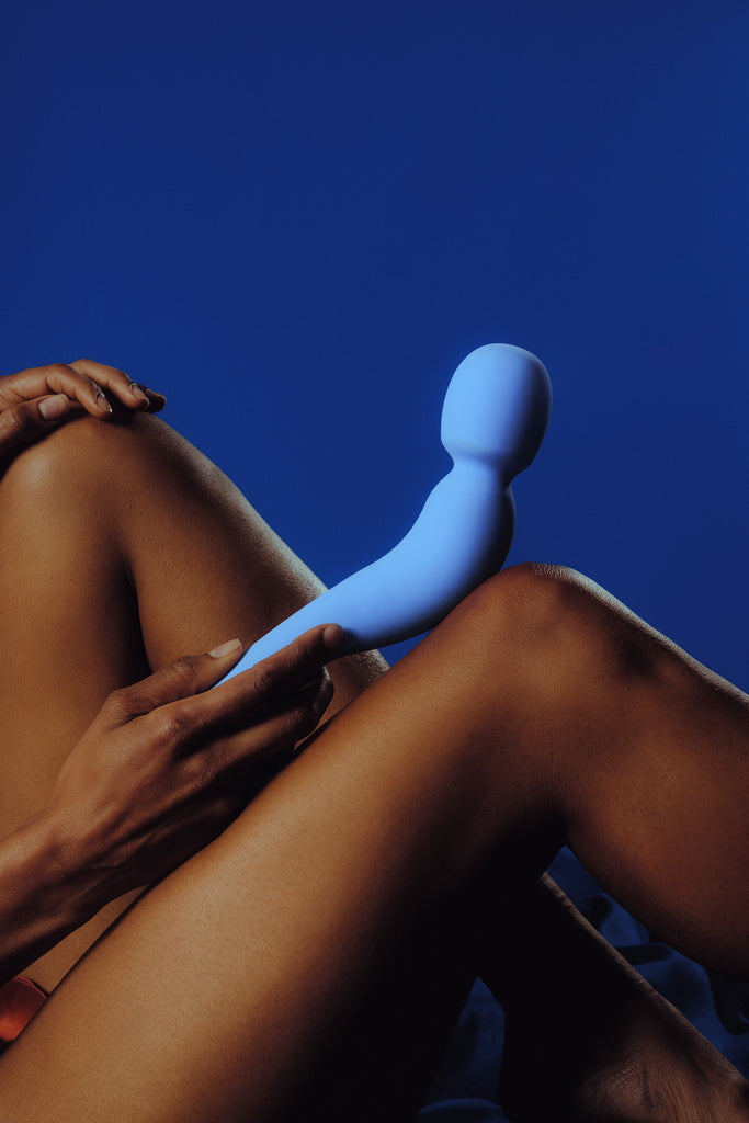women holding Dames Com wand vibrator displayed resting on legs in colour periwinkle in front of dark blue background.