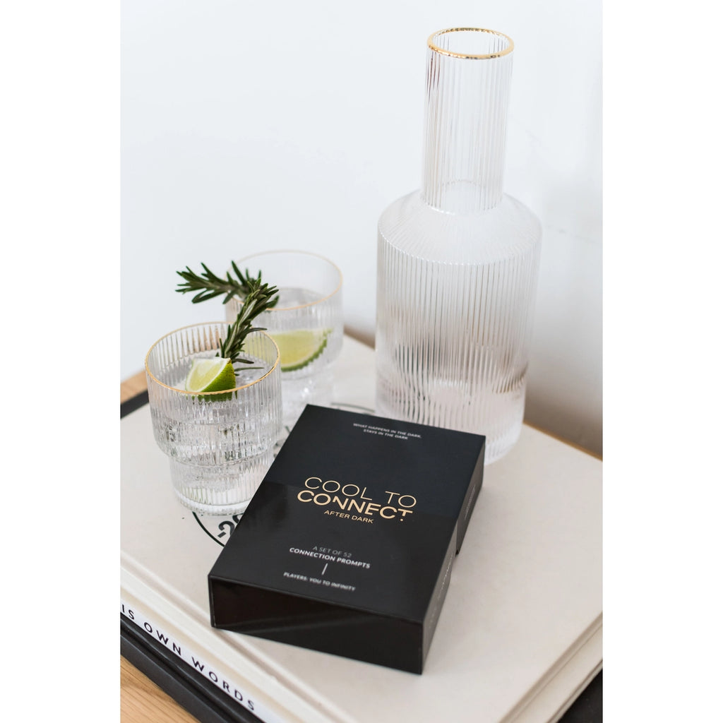 Cool To Connect After Dark edition displayed in lifestyle oriented scene sitting on stack of books. Includes large clear water carafe and gold rimmed glasses containing lime and rosemary.  