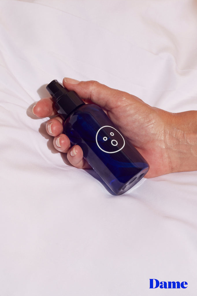 Dark blue bottle of alu lube in 118ml lightly held in a womans hands laid on solid white bedsheets with Dame logo in bottom right corner. 